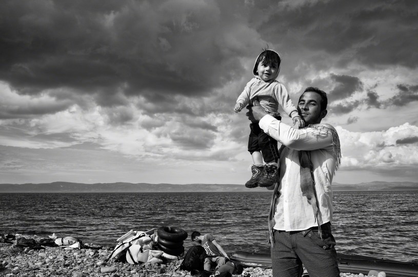 A black and white photograph of a father raising his young child to celebrate his family's safe passage to Lesbos after crossing over the Aegean Sea from Turkey